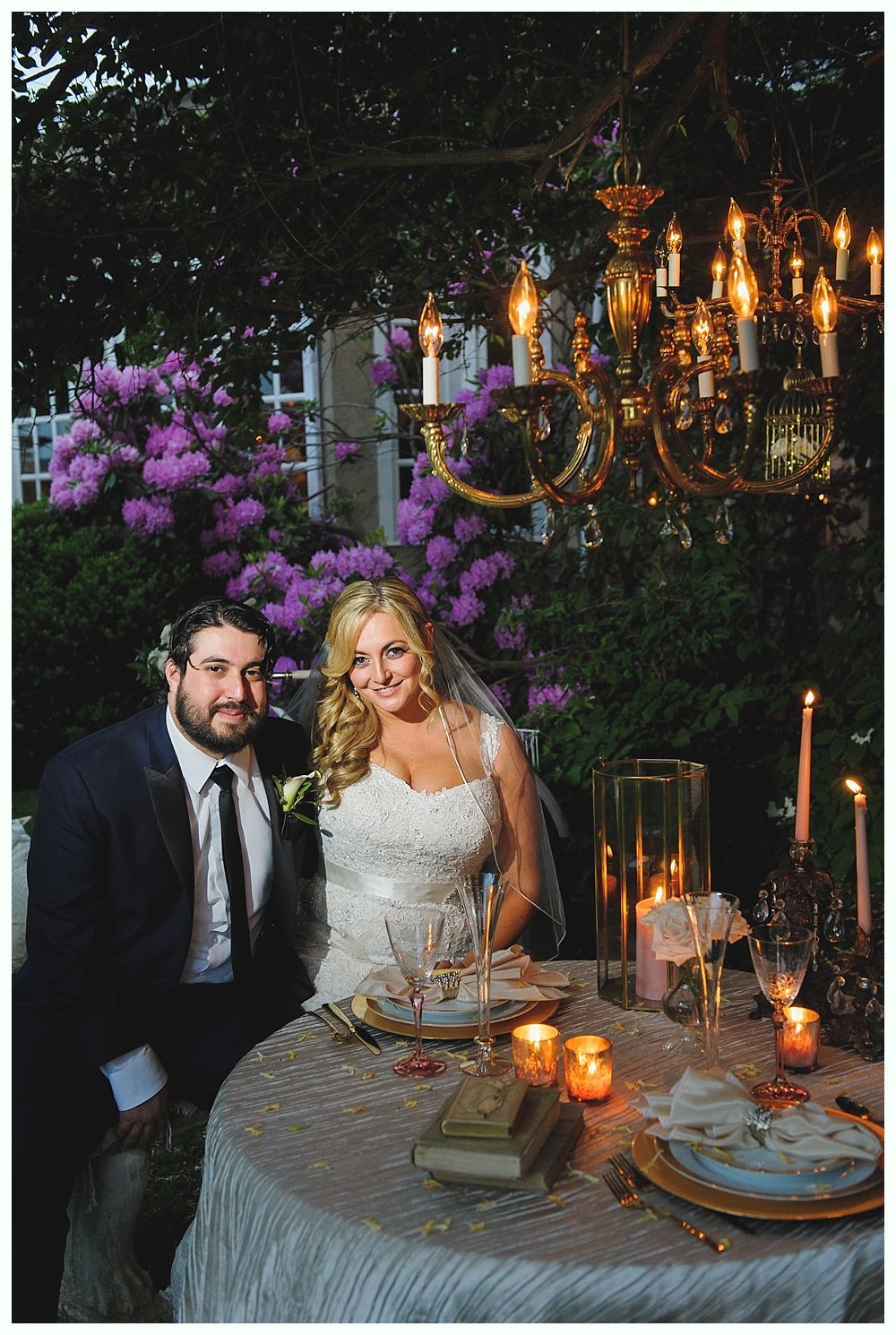 Bride and groom seated at a decorated table outdoors, under a chandelier, smiling.