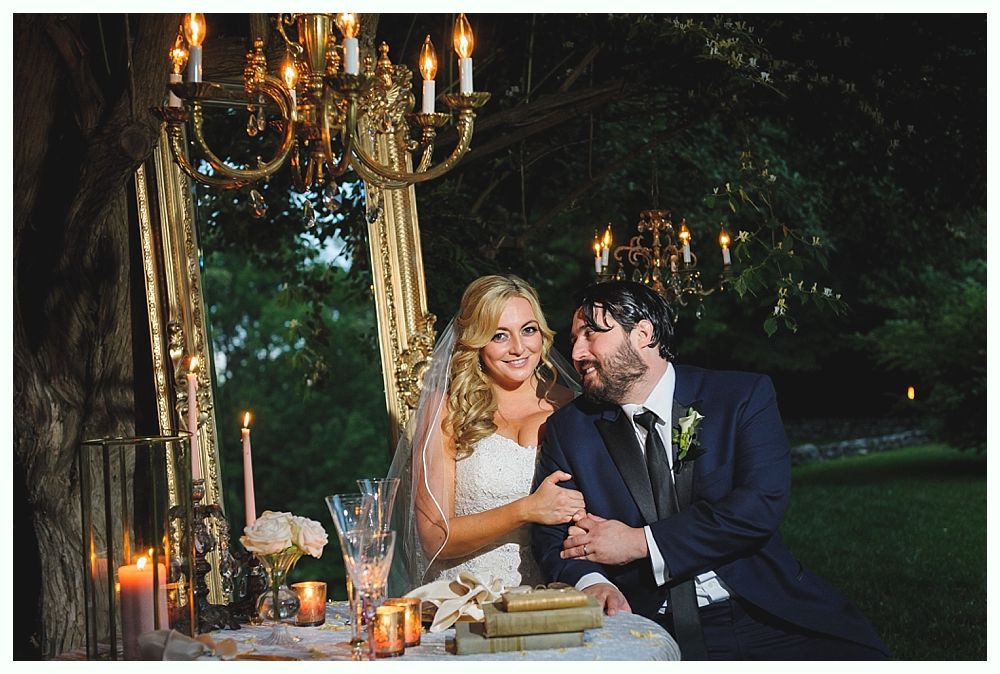 Bride and groom at a candlelit table beneath a gold chandelier. She smiles, he looks at her, both in formal wear.