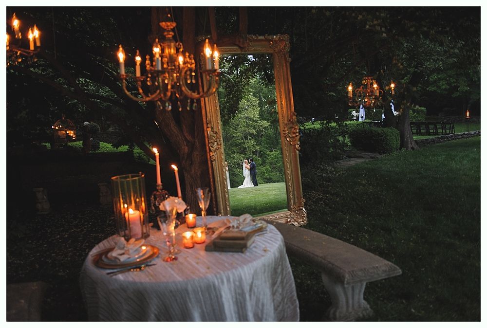 Outdoor wedding reception with table, candles, chandelier, and a mirror reflecting a couple on a lawn.