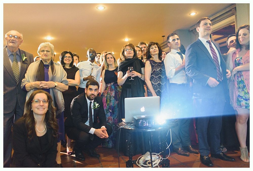 Group watches a laptop projection; a seated man kneels, some stand, looking up. Indoors, warm lighting.