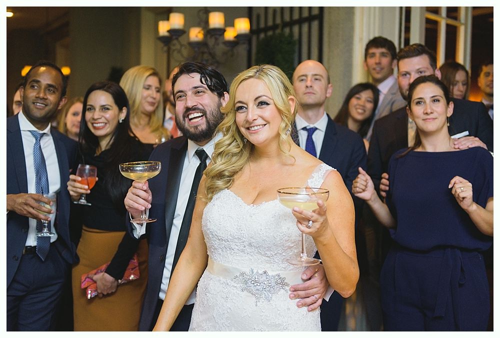 Bride and groom smiling, holding drinks, surrounded by guests at a wedding reception.