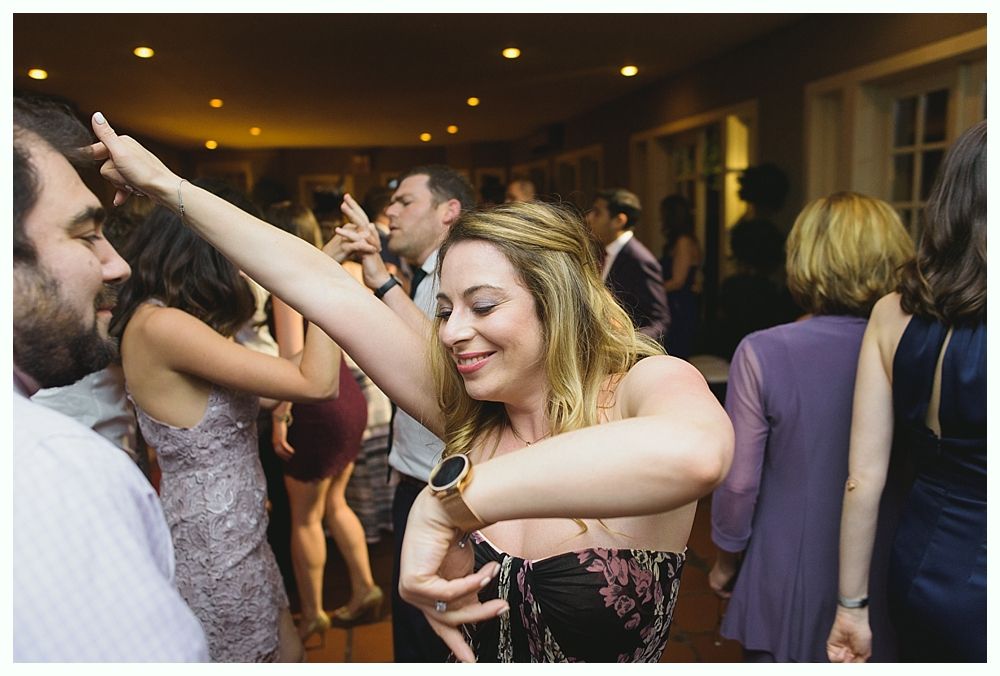 Woman dancing energetically at a party, arms raised. Many people in background, warm lighting.