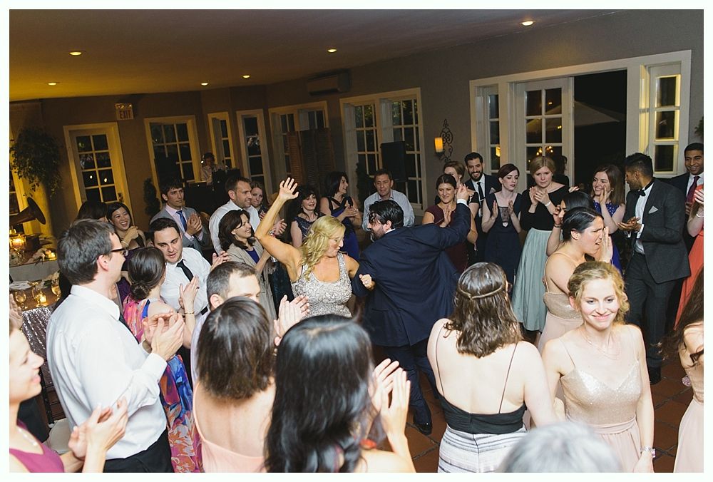 People dancing at an indoor event, clapping and celebrating. Windows line the walls; warm lighting.