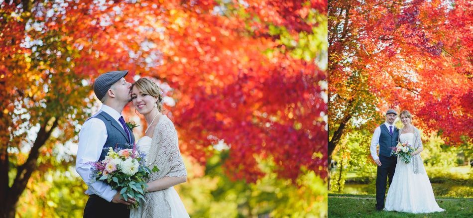 Couple kissing under vibrant red and yellow autumn tree. Groom in vest and hat, bride in white dress, holding flowers.