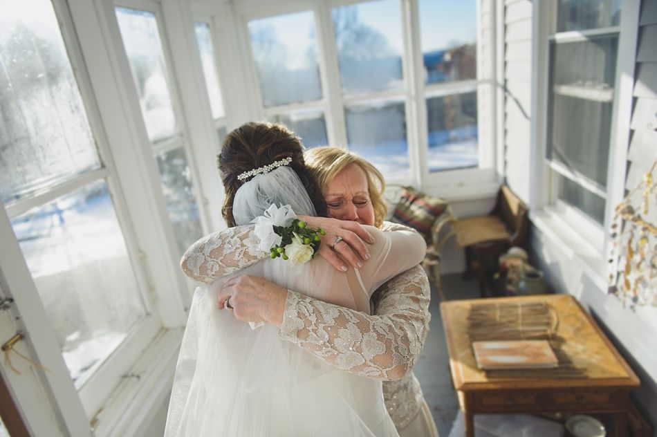 Bride and woman embrace in a sunlit room, windows in background. Woman in lace hugs bride wearing a veil and flowers.