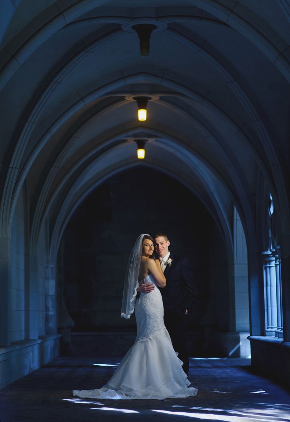 Bride and groom embrace in a stone archway. Bride wears a white mermaid gown, groom in a dark suit. Dark blue tones.