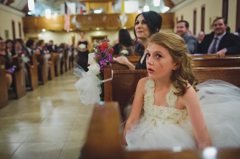 Flower girl in white dress looks up in a church pew. Others in pews watch the ceremony.