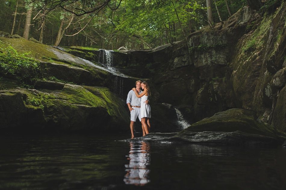 Couple kissing in water, standing near small waterfalls in a lush, green forest.