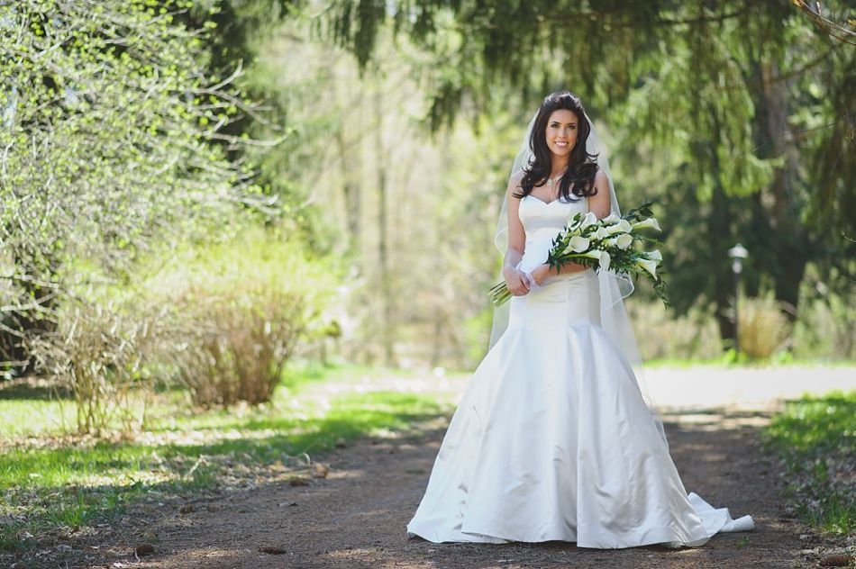 Bride in white dress holding flowers, standing on a path in a park, smiling.