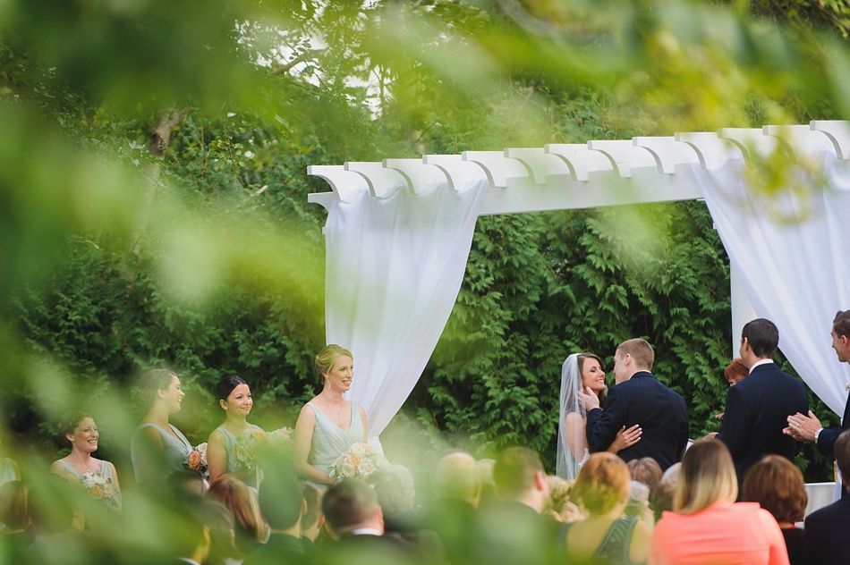 Wedding ceremony under white arbor; couple exchanging vows, surrounded by attendants and guests.