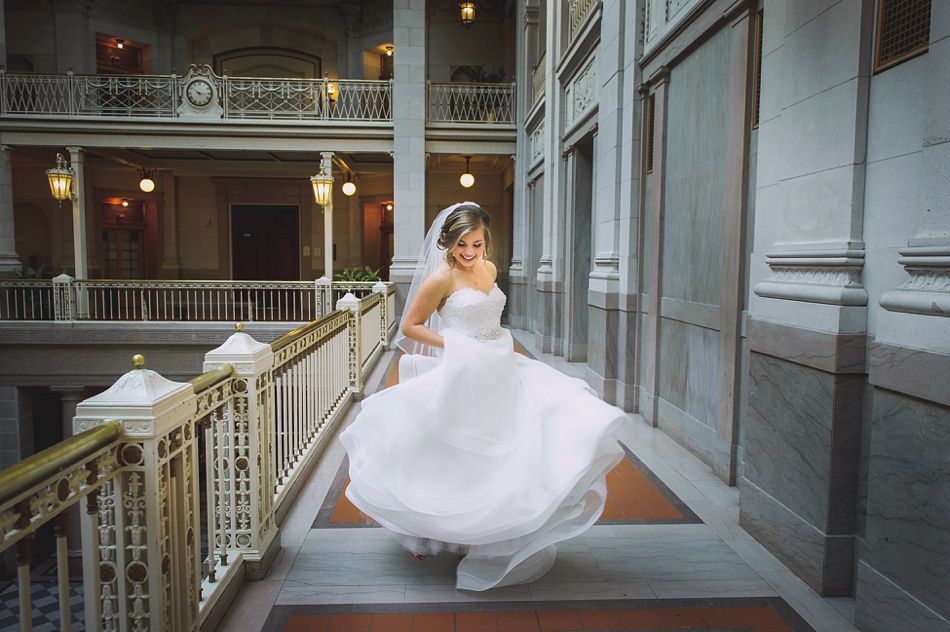 Bride twirls in a white wedding gown on a grand staircase, smiling. Interior shot with balconies and ornate railings.
