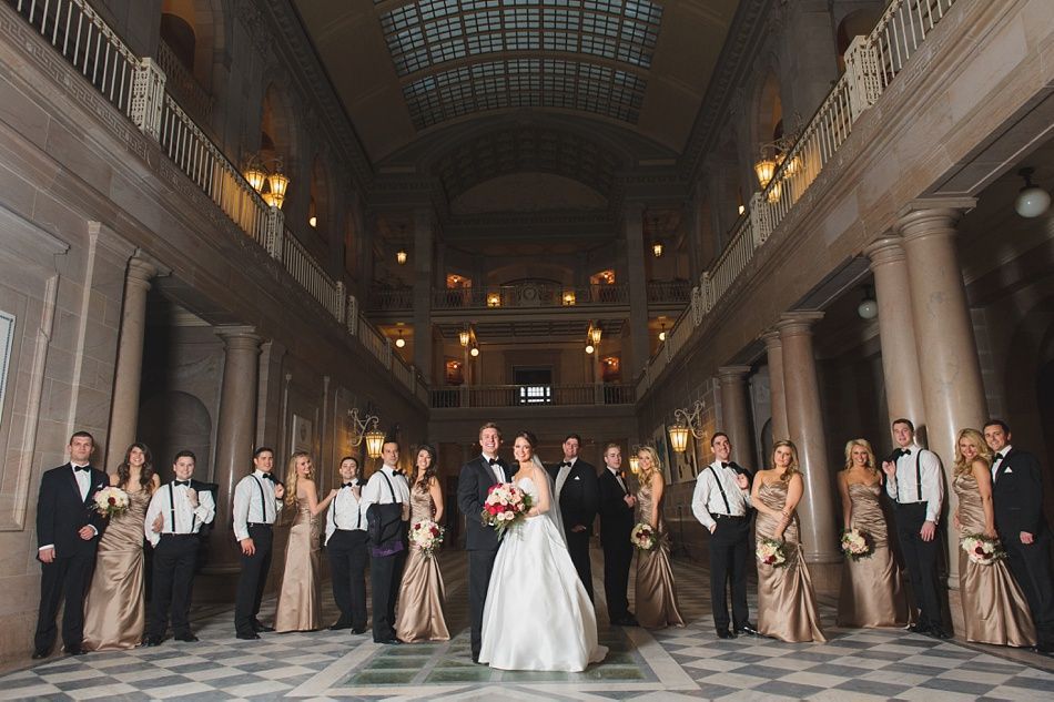 Wedding party posing in a grand hall with pillars, bride and groom centered, bridesmaids in gold dresses.