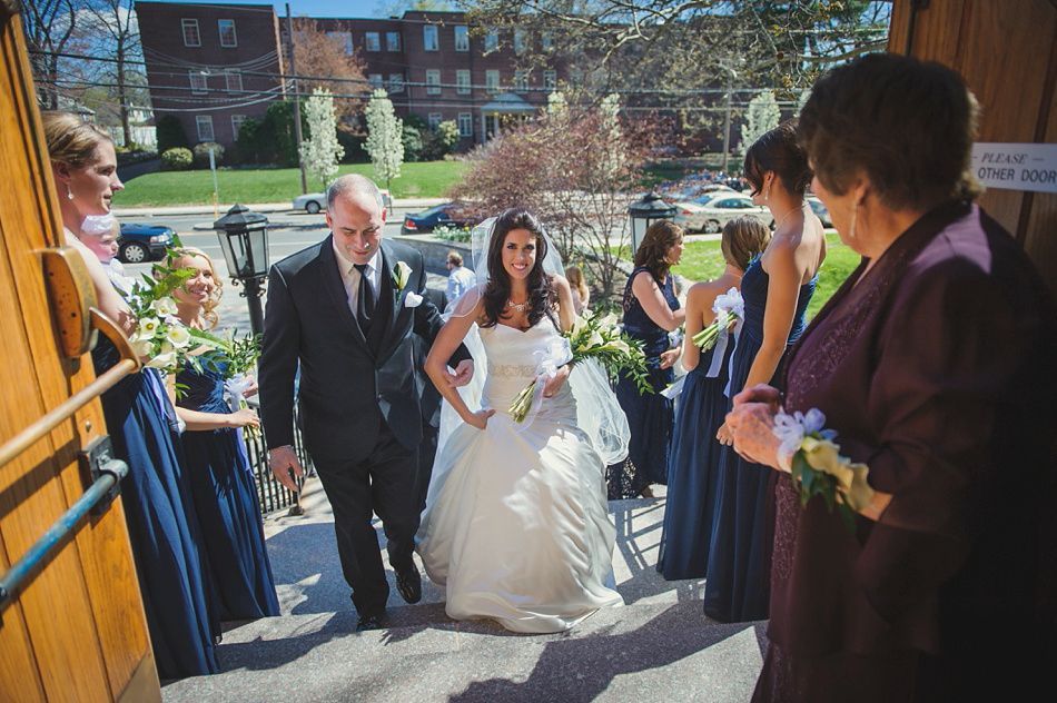 Bride in white gown exiting a church with her father. Bridesmaids in blue dresses are nearby. Sunny day.