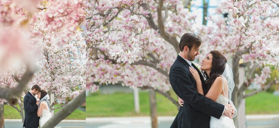 Couple embraces under a blossoming tree, faces close, in wedding attire. Pink and white flowers frame them.