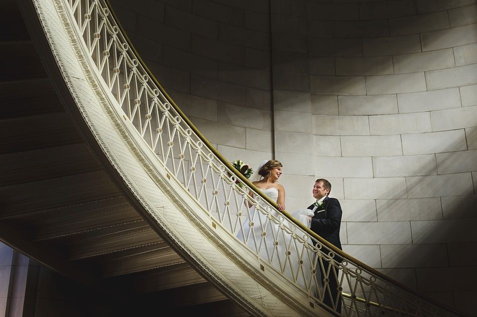 Bride and groom on a curved staircase, posing. Bride in white dress, groom in tuxedo. Stone wall, sunlight.