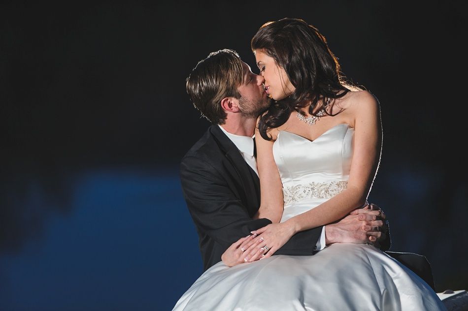 Bride and groom embrace in a kiss. Bride in white dress, groom in black suit, dark background.