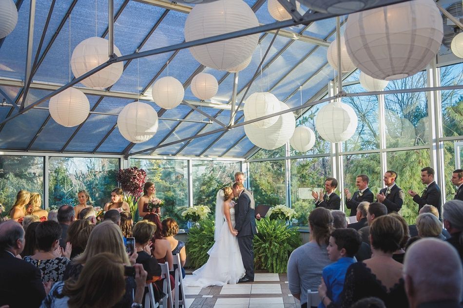 Bride and groom kissing during wedding ceremony in glass greenhouse, surrounded by guests. White lanterns hang from ceiling.