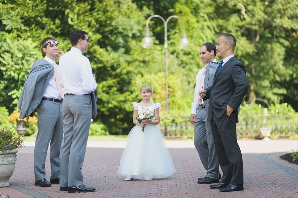Groomsmen and flower girl stand on a paved path outdoors. Men wear suits, flower girl in a white dress. Green trees in background.