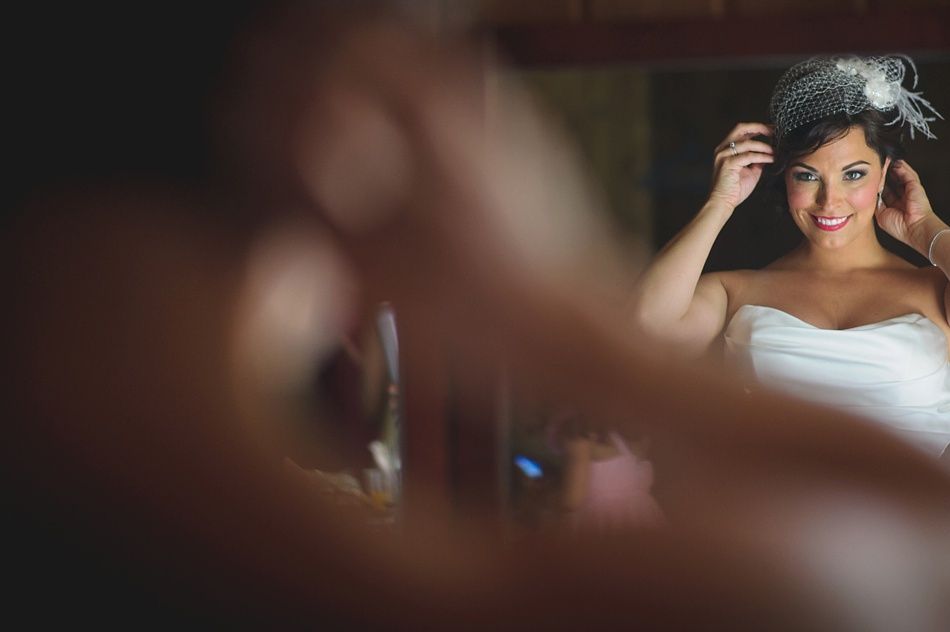 Bride in strapless dress, adjusting headpiece in mirror. Smiling, indoor setting with warm lighting.