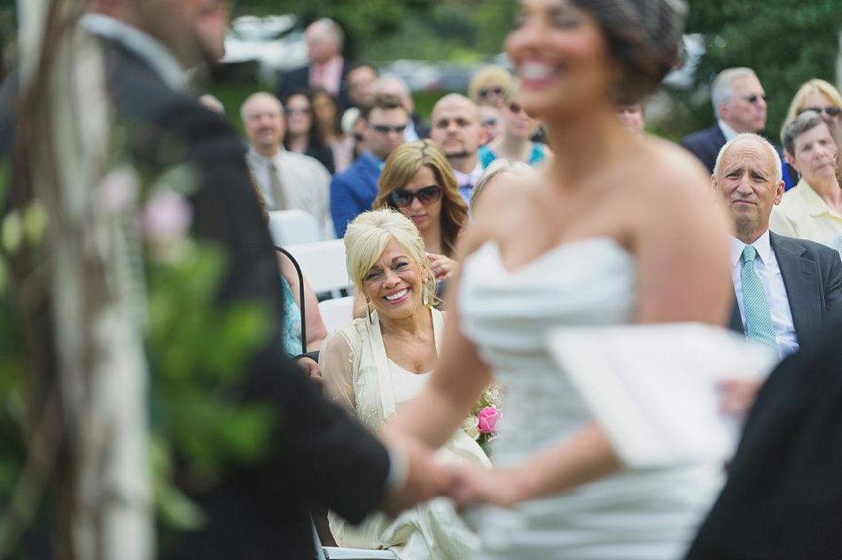 Wedding ceremony: Bride and groom holding hands, smiling. Guests watch, some smiling too. Outdoor setting.