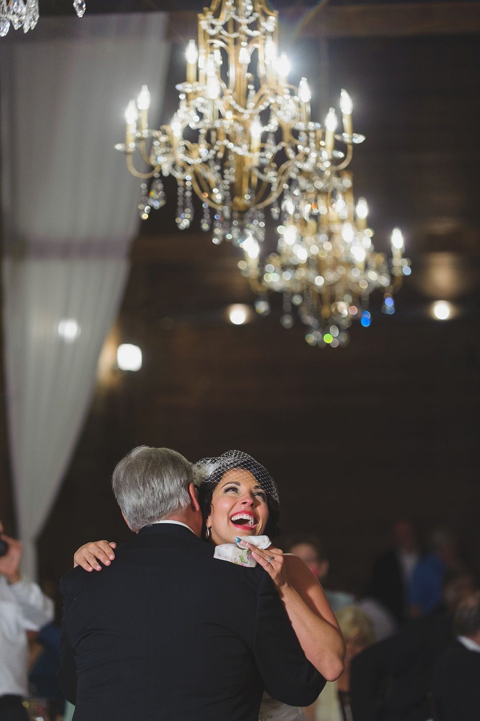 Bride dancing with a man under a chandelier; happy expressions in a dim reception hall.