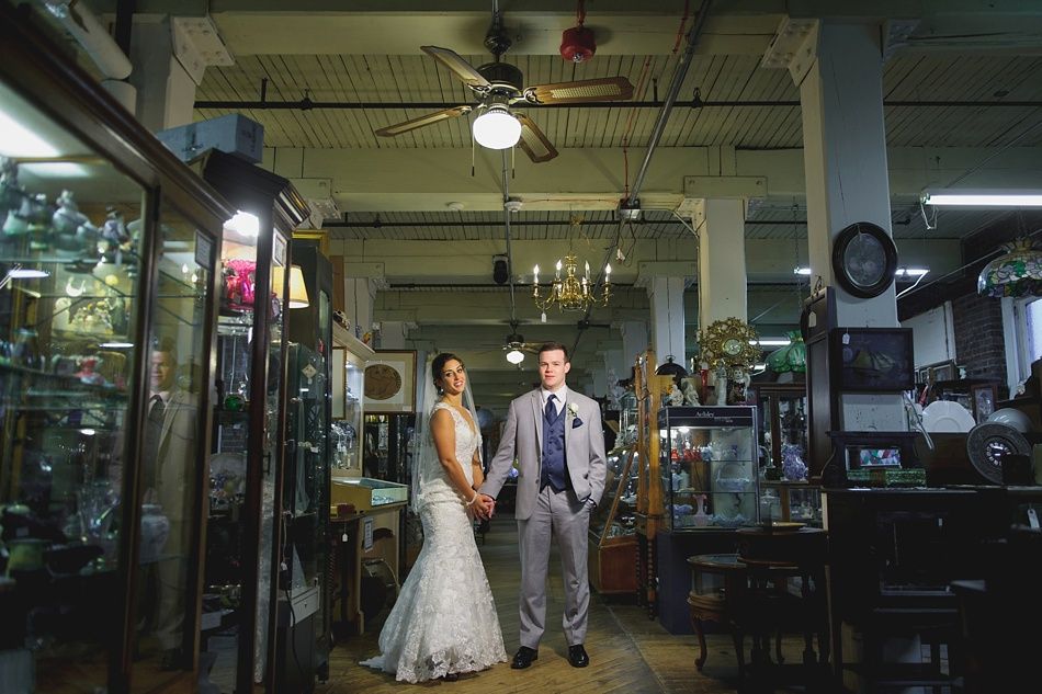 Bride and groom holding hands, posing in an antique shop. Bride wears white gown, groom wears gray suit.
