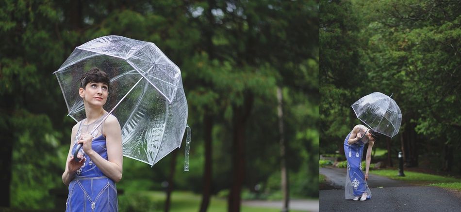 Woman with a clear umbrella in a blue dress, standing in the rain. Green trees in the background.