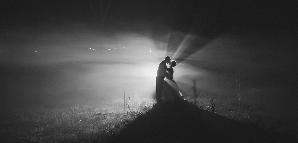 Couple embraces, illuminated by bright headlights in a foggy, dark field.