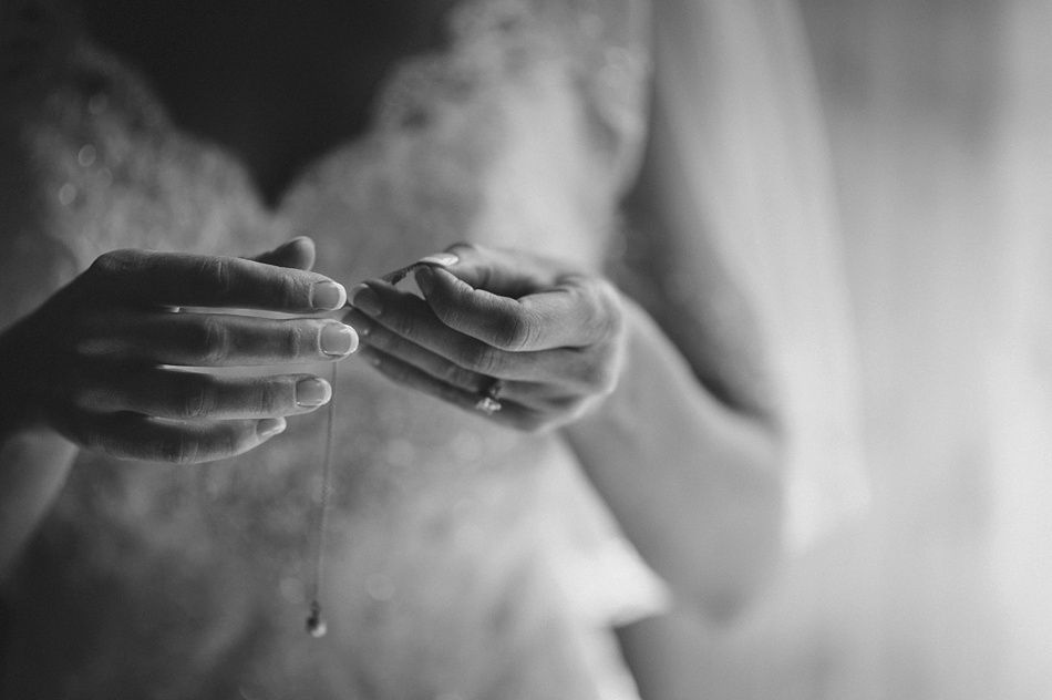 Bride's hands hold a delicate necklace, close up shot. She wears a lacy dress near a window.