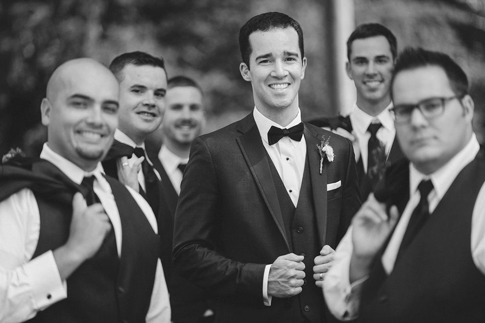 Groom in tuxedo with groomsmen, smiling. Black and white photo. Formal outdoor setting.