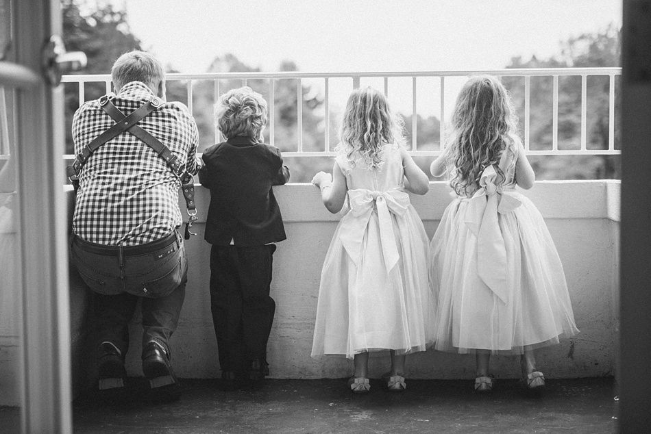 Children in formal attire look over a balcony railing; a boy in a suit, two girls in white dresses, and a person in a plaid shirt.