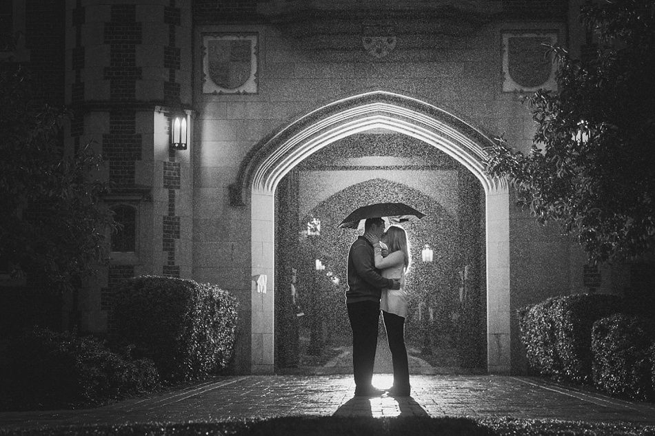 Couple embraces under umbrella in archway during rain at night.