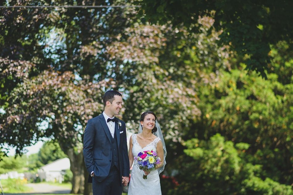 Bride and groom holding hands, smiling, walking outdoors near trees, with a colorful bouquet.