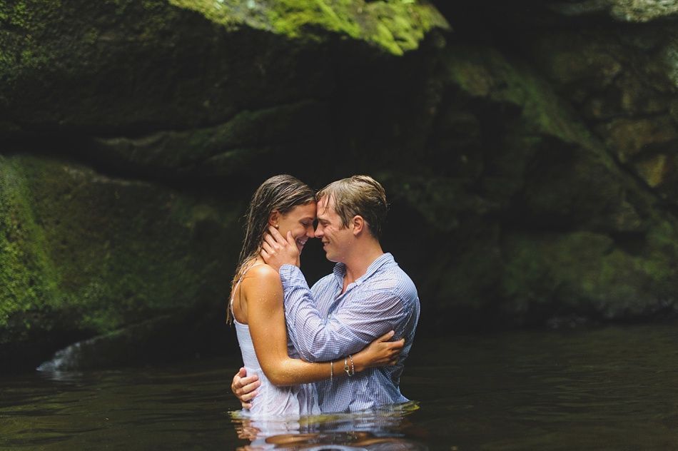 Couple embracing in a river, wet clothes, heads touching, dark water, lush green background.
