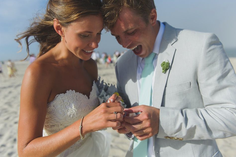 Bride and groom look at wedding rings on a sunny beach. The groom has an open mouth in excitement.