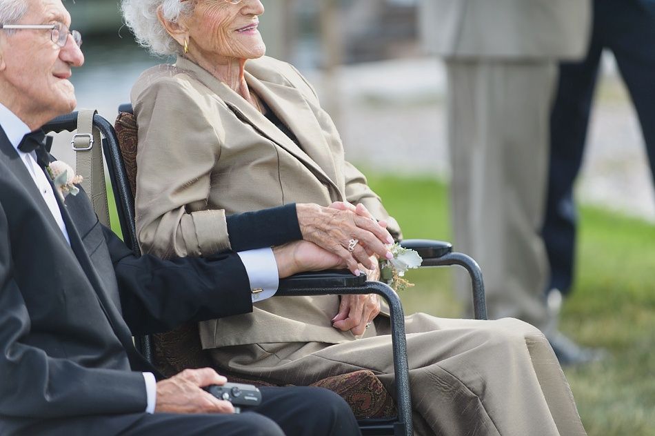 Elderly couple holding hands, the woman in a wheelchair, both dressed up outside.