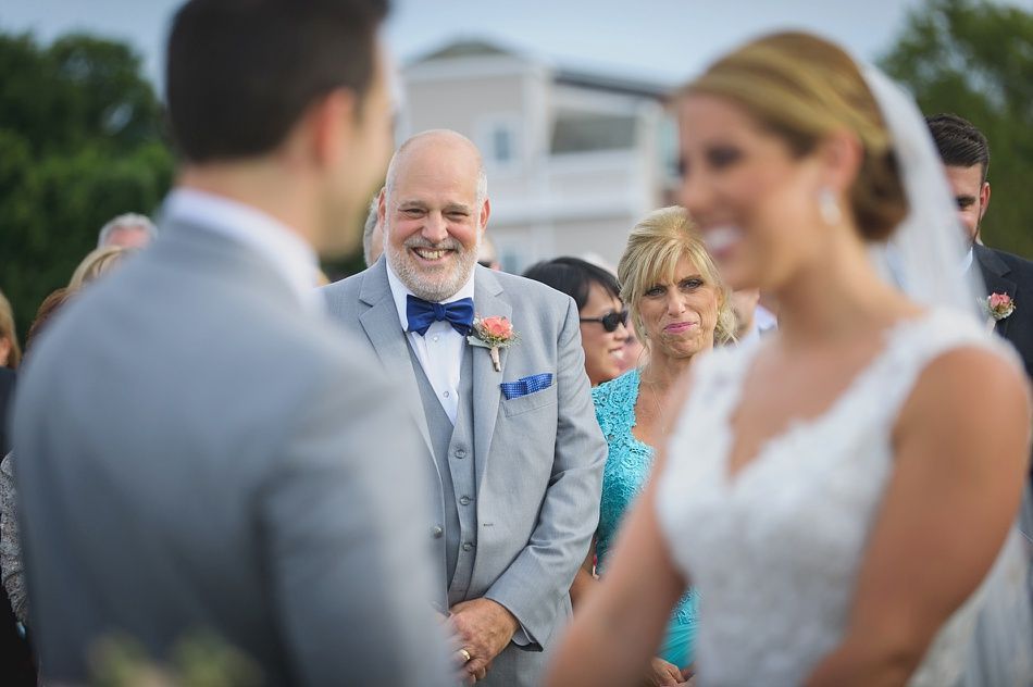 Wedding ceremony: Groom and bride face each other, father smiles, guests watch outdoors.