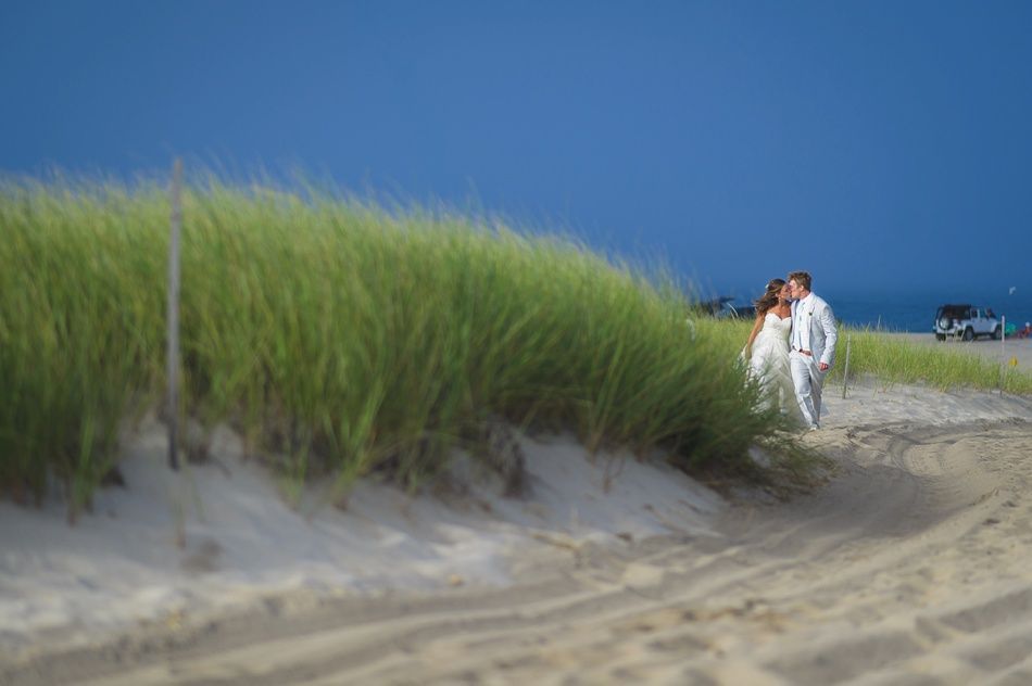 Couple in wedding attire walk along a sandy path, ocean and dark sky in background.