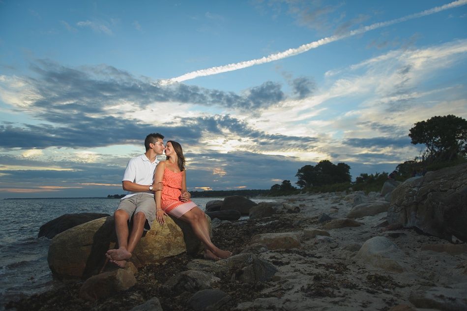 Couple sitting on rocks by water, about to kiss, under a cloudy sky at sunset.