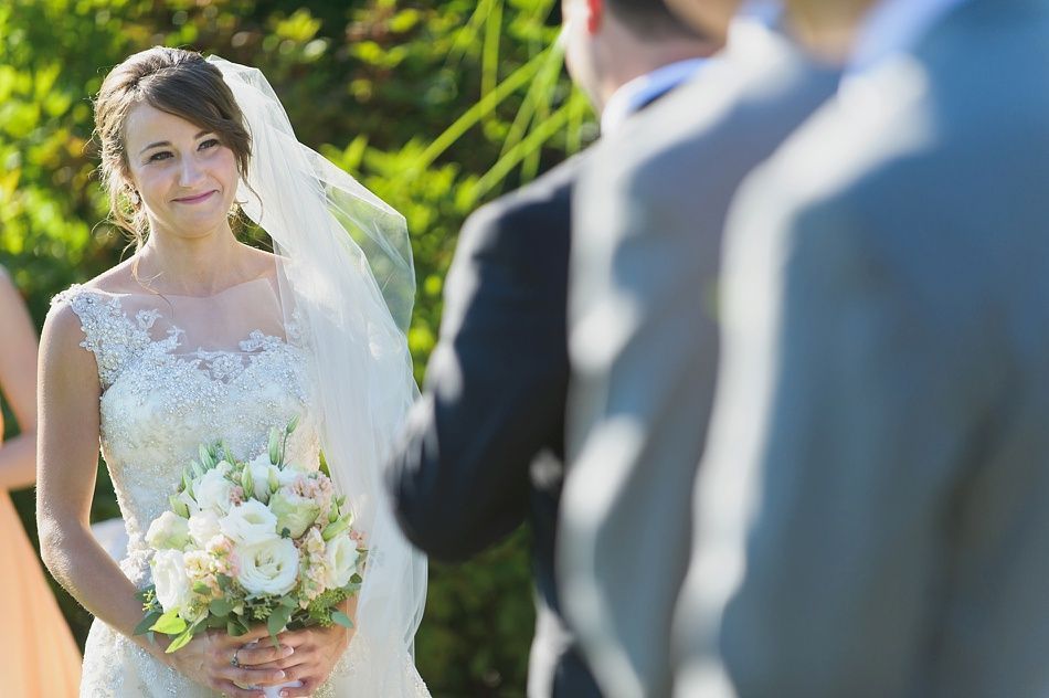 Bride in a lace gown, holding flowers, looks at groom during an outdoor wedding ceremony.