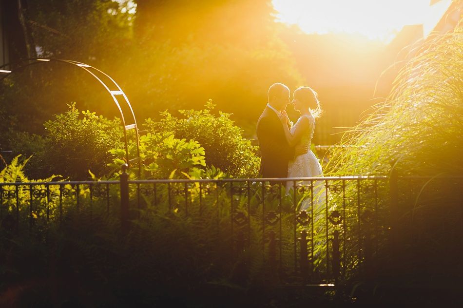 Couple embracing, bathed in golden sunlight, next to a wrought-iron fence and greenery.