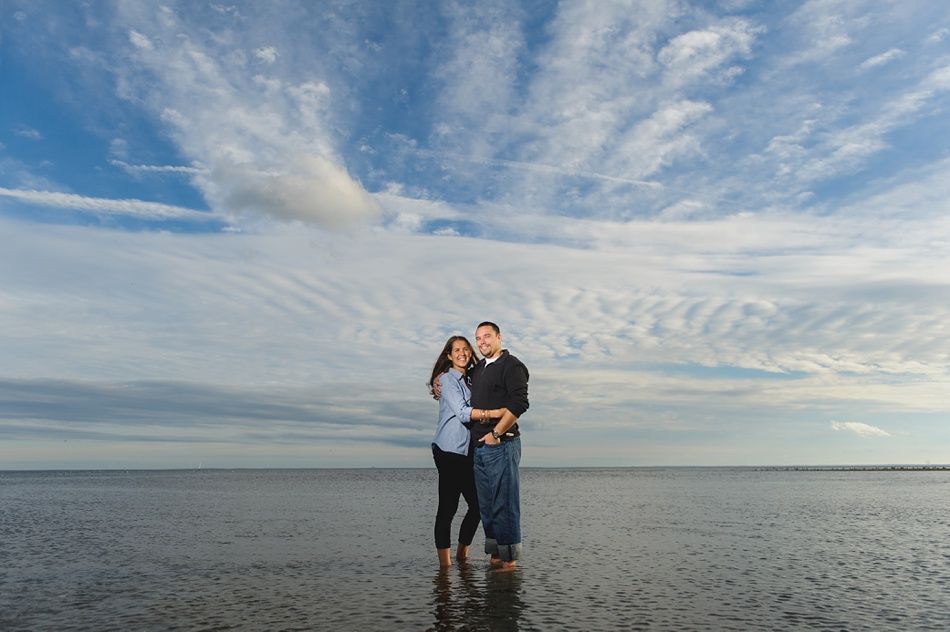 Couple standing, embracing at the beach. Blue sky with clouds overhead. They are smiling.