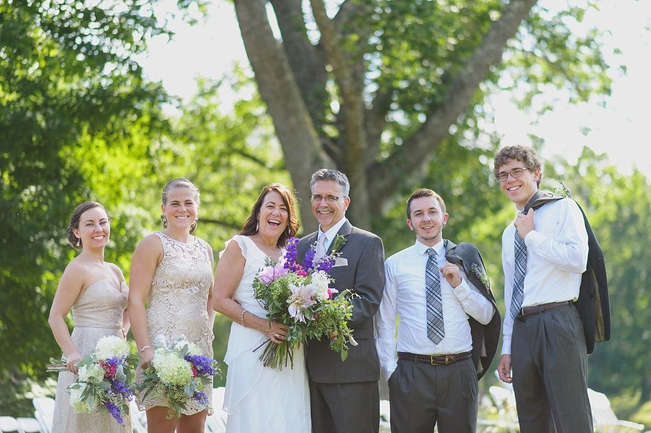Wedding party posing outdoors in front of a tree. Bride and groom smiling, bridesmaids, groomsmen. Sunny day.
