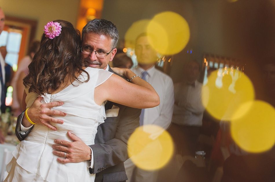 Bride in white dress hugs a man in a suit, both emotional, at a reception with blurred yellow lights.