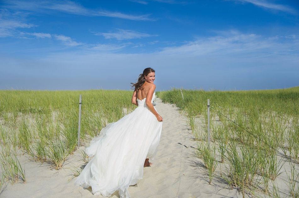 Bride running on sandy path toward camera, white dress flowing, blue sky and green grass.