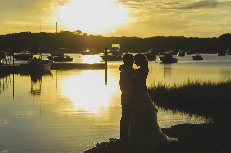 Couple silhouetted by sunset over water, embracing. Boats, dock, and trees in background.
