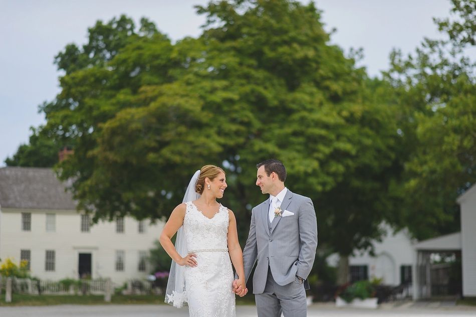 Bride and groom holding hands, smiling at each other. They stand outside a white building with trees in the background.