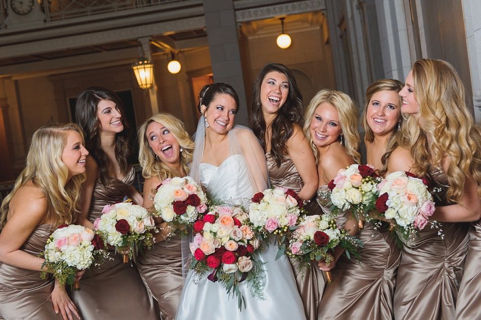 Bride and bridesmaids in gold dresses, laughing and holding bouquets, in a formal indoor setting.