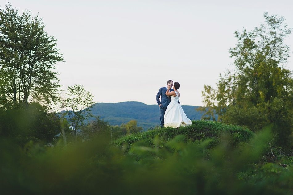 Couple embracing on a grassy hill; mountains in the background under a pale sky.
