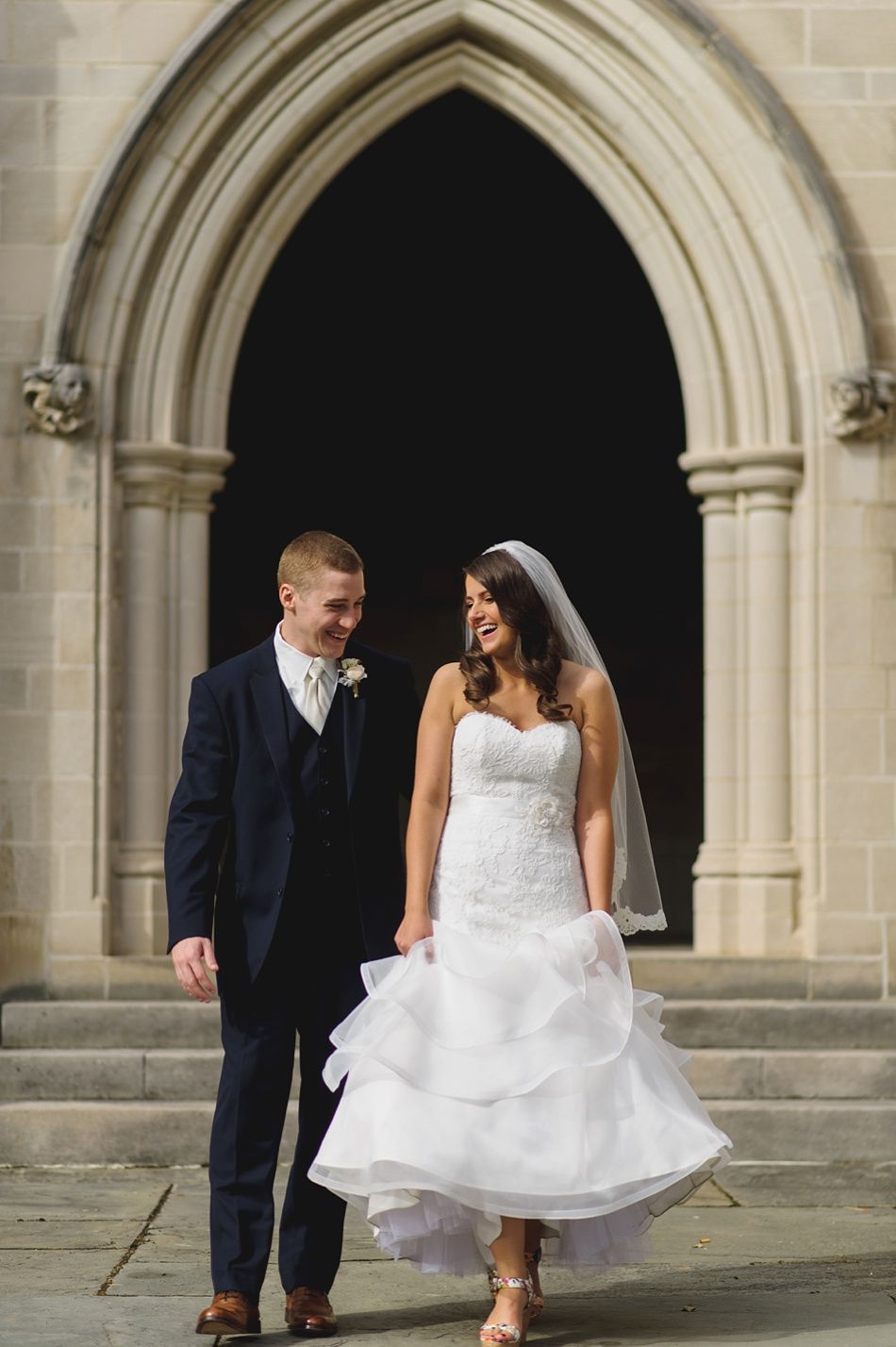 Bride and groom smiling, leaving church. Bride in white dress, groom in blue suit. Sunlight.